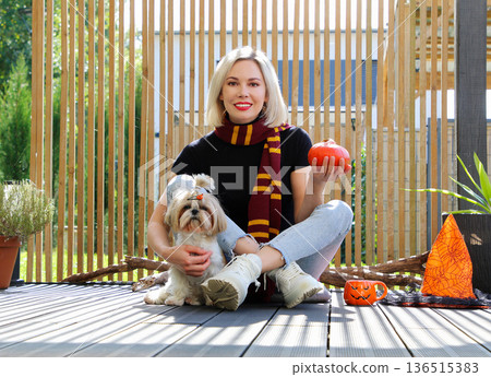 A beautiful girl is sitting on the terrace with a dog, holding a pumpkin in her hand. A beautiful girl is sitting on the terrace with a dog, holding a pumpkin in her hand. 136515383