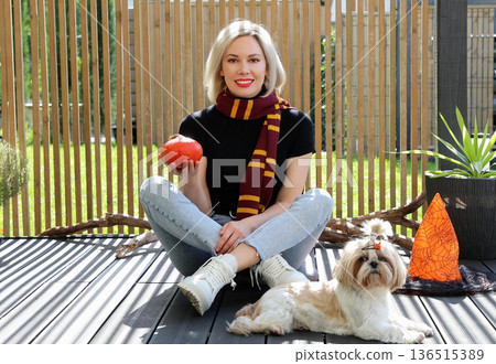A beautiful girl is sitting on the terrace with a dog, holding a pumpkin in her hand. A beautiful girl is sitting on the terrace with a dog, holding a pumpkin in her hand. 136515389