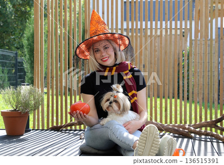 A beautiful girl is sitting on the terrace with a dog, holding a pumpkin in her hand. A beautiful girl is sitting on the terrace with a dog, holding a pumpkin in her hand. 136515391