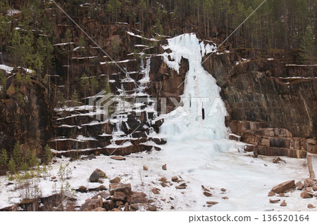 Large frozen waterfall cascading down a rocky cliff with surrounding snow and pine trees. Winter landscape at a cold quarry area. Granite quarry, Stolby Krasnoyarsk Large frozen waterfall cascading down a rocky cliff with surrounding snow and pine trees. Winter landscape at a cold quarry area. Granite quarry, Stolby Krasnoyarsk 136520164