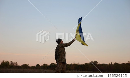 Male military waving blue-yellow banner on liberated territory occupied by russia. Young soldier of ukrainian army lifted flag of Ukraine in honor of victory against russian aggression at dusk 136524125
