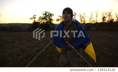 Soldier of ukrainian army going through meadow with Ukraine flag as symbol of victory against russian aggression. Young male military in uniform walking with blue-yellow banner at countryside Soldier of ukrainian army going through meadow with Ukraine flag as symbol of victory against russian aggression. Young male military in uniform walking with blue-yellow banner at countryside 136524126