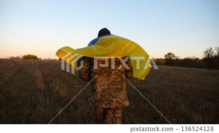 Young male military in uniform jogging with blue-yellow banner on shoulders at meadow. Soldier of ukrainian army running with flag of Ukraine as symbol of victory against russian aggression. Slow mo 136524128