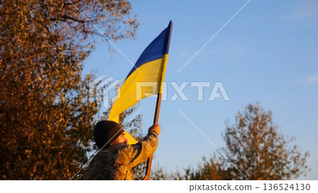 Male military in uniform waving flag of Ukraine at countryside. Young soldier of ukrainian army lifting blue-yellow banner as symbol of victory against russian aggression. Invasion resistance concept 136524130