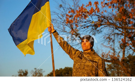 Male ukrainian army soldier lifting blue-yellow banner in honor of victory against russian aggression. Young military in camouflage uniform raised flag of Ukraine at countryside. Invasion resistance 136524162