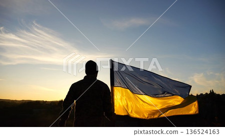 Silhouette of ukrainian army man stands with national banner at countryside on sunset. Male soldier in military uniform with Ukraine flag as symbol of victory against russian aggression. Slow motion Silhouette of ukrainian army man stands with national banner at countryside on sunset. Male soldier in military uniform with Ukraine flag as symbol of victory against russian aggression. Slow motion 136524163