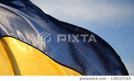 Blue-yellow symbol of Ukraine waving at wind against the background of sunlight. Ukrainian flag illuminated by sun fluttering at countryside. Concept of peace and support against russian aggression 136524165