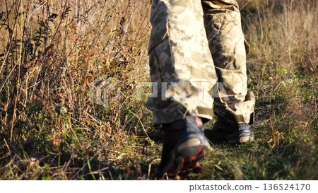 Low view to male feet of ukrainian soldiers going along trail at countryside. Legs of young army man walking through dry grass at sunny day. War in Ukraine. Slow motion Low view to male feet of ukrainian soldiers going along trail at countryside. Legs of young army man walking through dry grass at sunny day. War in Ukraine. Slow motion 136524170