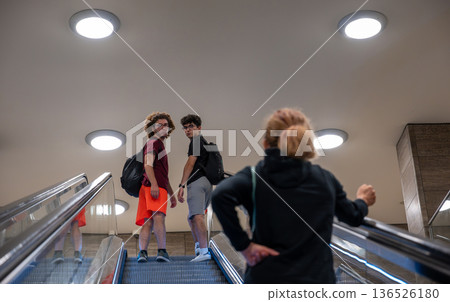 Berlin, germany, august 13, 2023. Three people riding an escalator in a modern urban indoor setting. Two young men are looking back from an ascending escalator 136526180