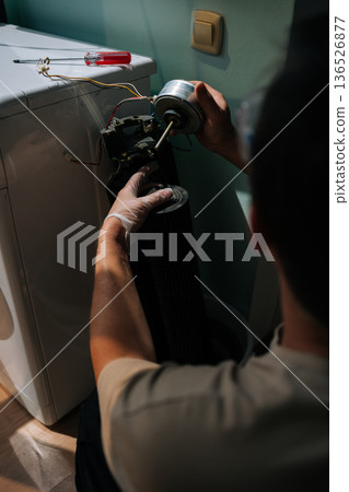 Rear view of unrecognizable technician male wearing safety gloves examines components of disassembled washing machine, focusing on detached motor while troubleshooting malfunction. 136526877