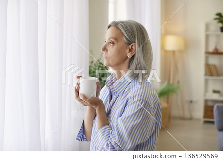 Senior Woman Daydreaming By Window With Coffee Cup At Home 136529569