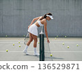 Female tennis player in white sportswear picking up ball on modern outdoor court during training 136529871