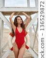 Young woman in red swimsuit posing by lifeguard tower on sandy beach with ocean backdrop 136529872
