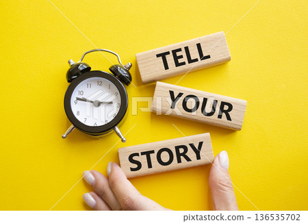 Tell your story symbol. Wooden blocks with words Tell your story. Businessman hand. Beautiful yellow background with alarm clock. Business and Tell your story concept. Copy space. 136535702