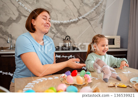 Smiling mother and happy little girl play together with rabbit glove, sitting at Easter table. Spring holiday 136539921
