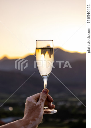 Man holding champagne glass with vitiligo in hand, sunset over Stellenbosch mountains. Celebration of fine wine in Cape Winelands. Man holding champagne glass with vitiligo in hand, sunset over Stellenbosch mountains. Celebration of fine wine in Cape Winelands. 136540421