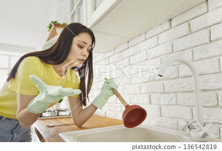Woman wearing rubber gloves holding plunger stands in cozy kitchen beside clogged sink 136542819