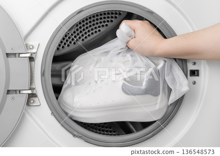 A woman's hand places white sports sneakers into a special laundry bag in the washing machine A woman's hand places white sports sneakers into a special laundry bag in the washing machine 136548573