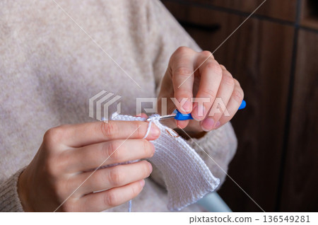 Female artisan with light skin using a blue crochet hook to create a white fabric piece while seated indoors with wooden background elements visible 136549281