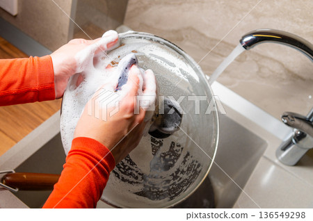 Female Person with orange long-sleeve shirt scrubbing a glass pot with soap under running water in a modern kitchen sink with a stylish faucet and countertop Female Person with orange long-sleeve shirt scrubbing a glass pot with soap under running water in a modern kitchen sink with a stylish faucet and countertop 136549298
