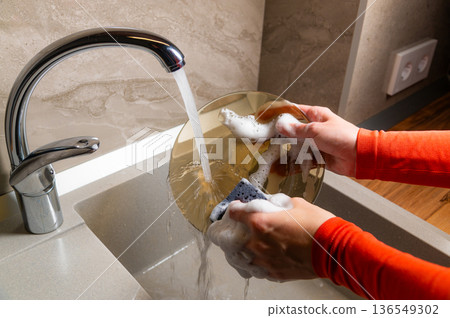 Person with orange long-sleeve shirt washes a round golden plate under running water in a modern kitchen sink with a sleek faucet and textured wall 136549302