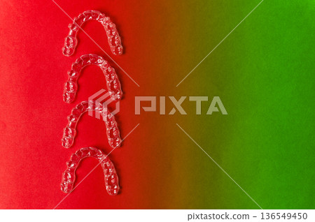 Transparent dental aligners arranged in a row on a gradient background of red and green, showcasing orthodontic treatment options for teeth straightening 136549450