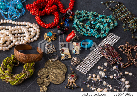 Collection of vintage and bohemian jewelry, featuring turquoise beads, pearl necklaces, coral pieces, silver rings, and brass bracelets scattered on a dark textured background 136549693