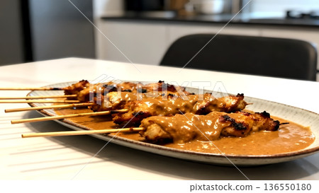 Grilled chicken satay skewers topped with creamy peanut sauce, served on a ceramic plate in a modern kitchen. 136550180