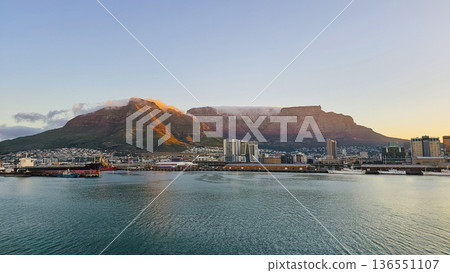 Panoramic view of Cape Town harbor with turquoise water, gentle ripples, diverse docked ships, urban skyline, and Table Mountain capped with clouds 136551107