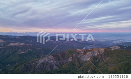 Panoramic view of Georgian mountain ranges at pre-dawn or sunset with forested slopes, rocky ridges, pastel sky, distant snow peaks, and small village lights Panoramic view of Georgian mountain ranges at pre-dawn or sunset with forested slopes, rocky ridges, pastel sky, distant snow peaks, and small village lights 136551116