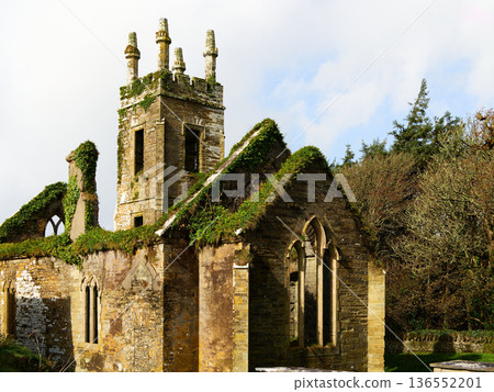 Old church ruins with green plants against a cloudy sky. Moss grows on an abandoned buildings roof. Exterior of a weathered stone church. Picturesque ruins of a historic chapel. 136552201