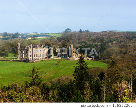 Castle, a stone ruin, stands on a vibrant green hillside under a clear blue sky. Dense forest covers distant hills, while farm buildings are visible in the background. Castle, a stone ruin, stands on a vibrant green hillside under a clear blue sky. Dense forest covers distant hills, while farm buildings are visible in the background. 136552203