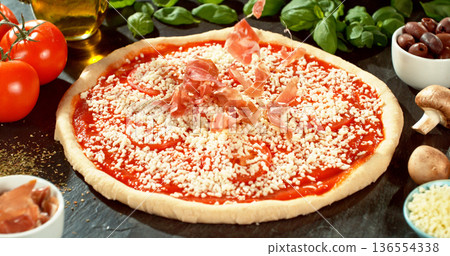 Fresh ham slices falling onto prepared pizza dough with tomato sauce and mozzarella. Italian pizza preparation on rustic wooden table. 136554338