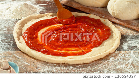 Closeup of spreading tomato sauce on fresh pizza dough. Wooden spoon moves gently across the surface, highlighting texture, freshness and authentic Italian pizza preparation. 136554357