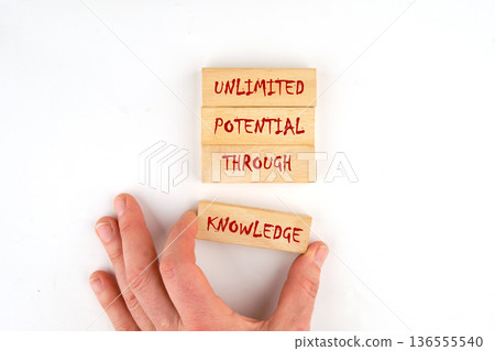 A hand places a wooden block labeled Knowledge with other blocks saying Unlimited Potential Through on a plain white surface 136555540