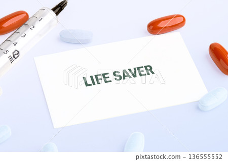 A white card proclaiming LIFE SAVER is centered among a 5ml syringe, several orange capsules, and white pills, displayed on a clean white surface A white card proclaiming LIFE SAVER is centered among a 5ml syringe, several orange capsules, and white pills, displayed on a clean white surface 136555552