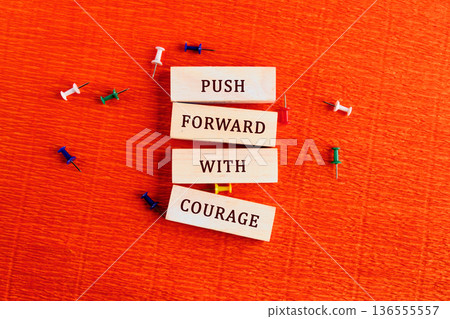 Wooden blocks are vertically stacked displaying the phrase Push Forward With Courage. Colorful pushpins are scattered on an orange textured surface 136555557
