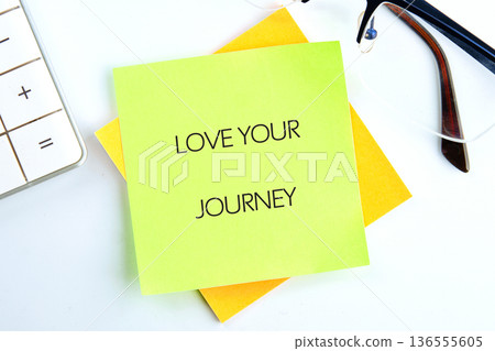 A sticky note with the text love your journey sits on a desk next to a calculator and a pair of glasses, showing an organized workspace A sticky note with the text love your journey sits on a desk next to a calculator and a pair of glasses, showing an organized workspace 136555605
