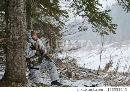 Soldier In Winter Camouflage Kneeling Against Tree With Rifle On Forest Patrol 136555643