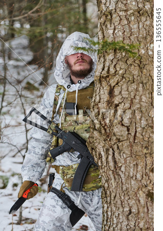 Armed Soldier In Winter Camouflage Peeking From Behind Tree With Rifle And Knife In Snowy Forest 136555645