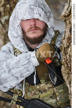 Winter Camouflaged Soldier With Knife and Tactical Vest Concealed Behind Tree in Snowy Forest 136555681