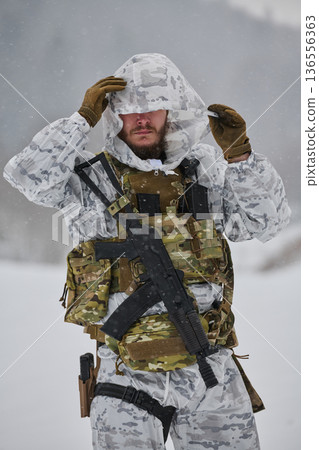 Winter Soldier In White Snow Camouflage Gear Adjusting Hood With Tactical Rifle And Vest 136556363