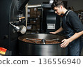 Male worker in an apron standing at the drum of an industrial coffee machine and controlling the process of cooling  136556940