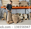 Warehouse worker folding a bag with coffee. Blue-collar worker managing bag at shelves in a warehouse. 136556947