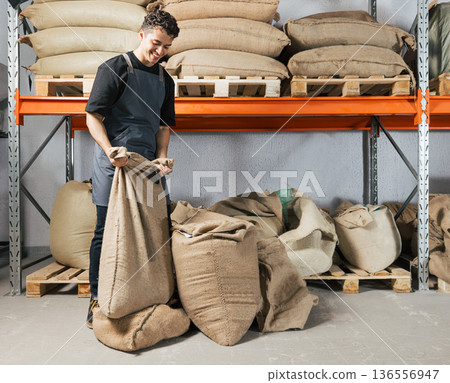 Warehouse worker folding a bag with coffee. Blue-collar worker managing bag at shelves in a warehouse. 136556947
