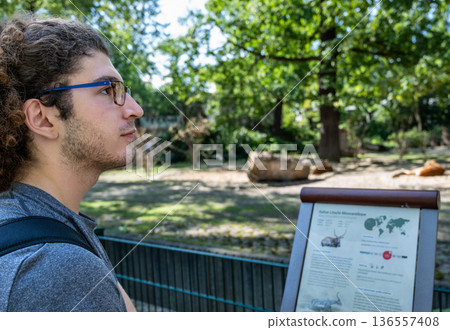 Berlin, germany, august 14, 2023. Young man with glasses reads an information board at berlin zoo, learning about native habitats, wildlife and conservation outdoors 136557408
