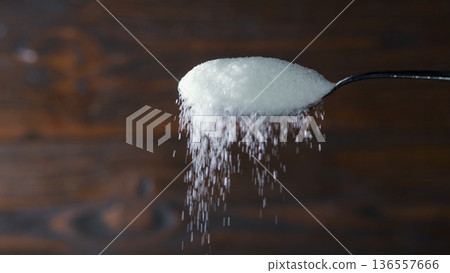 Sugar falls from a spoon onto a wooden surface in a simple kitchen setting Sugar falls from a spoon onto a wooden surface in a simple kitchen setting 136557666