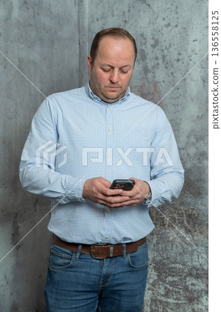 Man in blue shirt focused on phone screen indoors. Modern communication concept, remote work, texting, browsing internet and everyday technology use Man in blue shirt focused on phone screen indoors. Modern communication concept, remote work, texting, browsing internet and everyday technology use 136558125