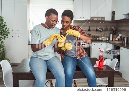 Two women wearing gloves using a smartphone while taking a break from cleaning the kitchen Two women wearing gloves using a smartphone while taking a break from cleaning the kitchen 136559096