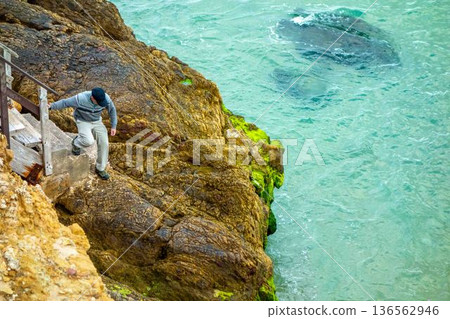 Man climbs onto cliff rocks at base of hiking trail steps by ocean Salema Beach Algarve Portugal Man climbs onto cliff rocks at base of hiking trail steps by ocean Salema Beach Algarve Portugal 136562946